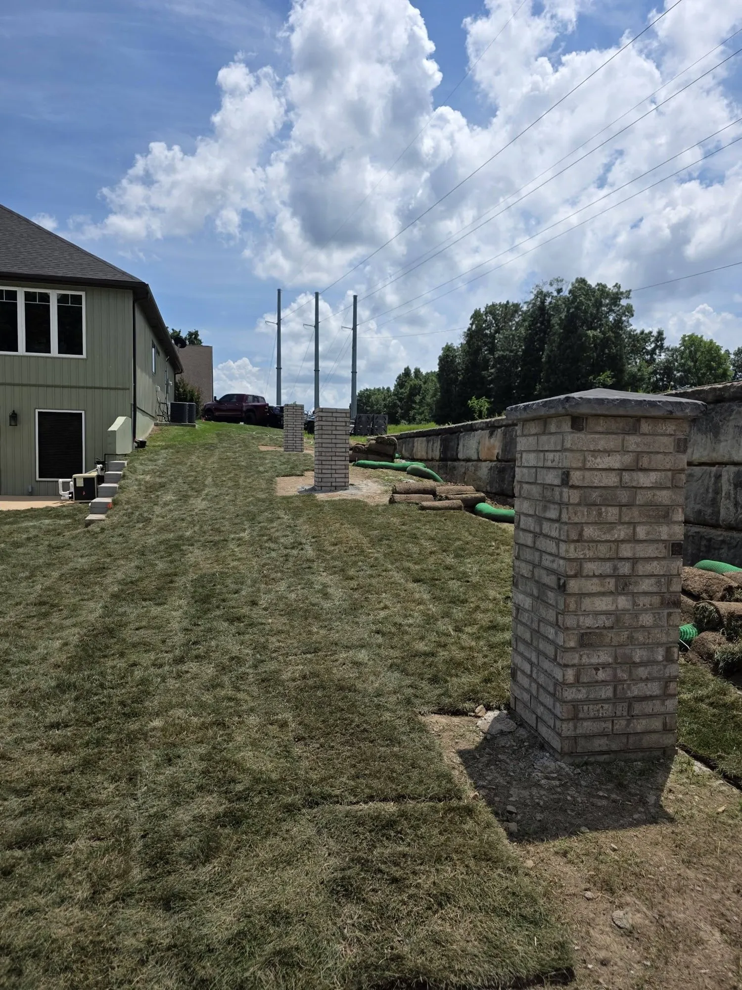 Row of new brick pillars with stone caps along a property line beside fresh sod and a large block wall
