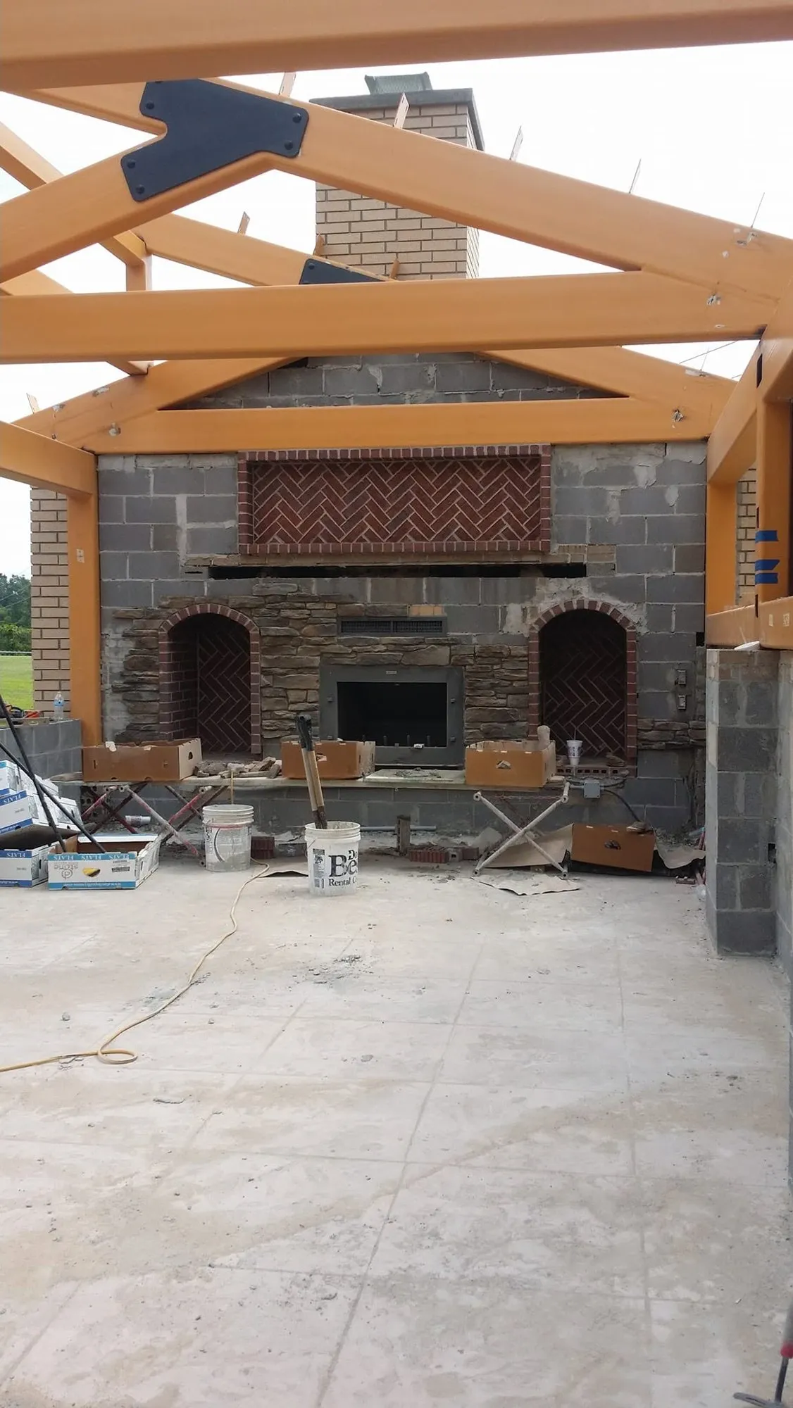 Outdoor pavilion fireplace under construction with stacked stone, arched wood niches and herringbone brick panels
